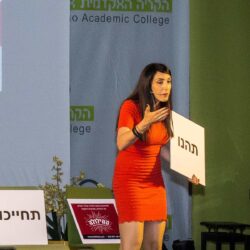 הילה עשור על הבמה עם שלט במהלך הרצאה
