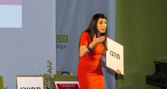 הילה עשור על הבמה עם שלט במהלך הרצאה