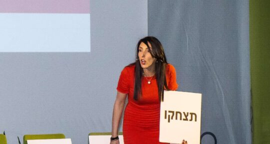 הילה עשור מרצה על הבמה עם שלט "תצחקו"