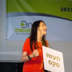 הילה עשור על הבמה עם שלט "תעשו סקס" בהרצאה לנשים