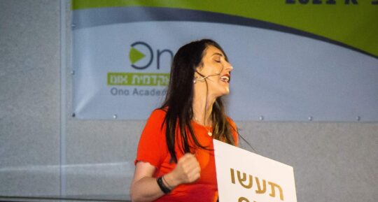 הילה עשור על הבמה עם שלט "תעשו סקס" בהרצאה לנשים