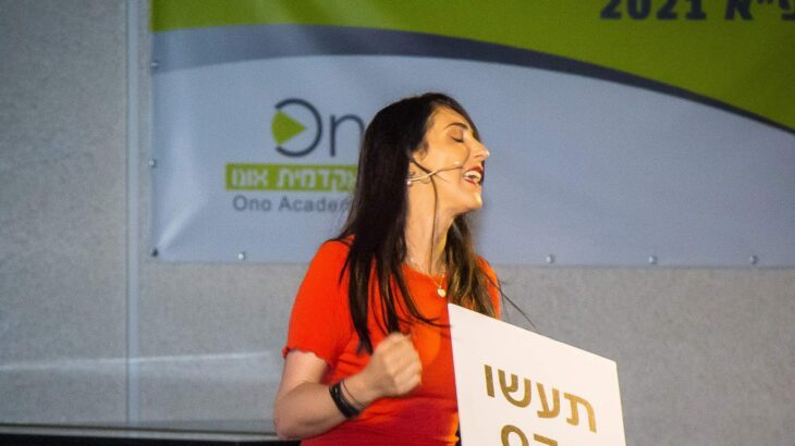 הילה עשור על הבמה עם שלט הומוריסטי בערב לנשים
