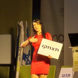 הילה עשור על הבמה עם שלט "תצחקו"