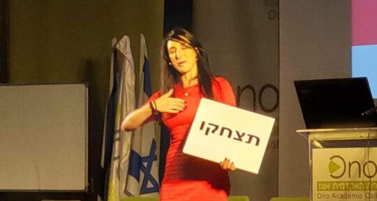 הילה עשור על הבמה עם שלט "תצחקו"